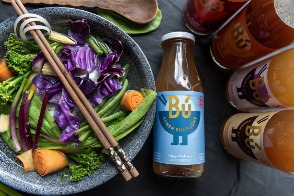 BU ramen broth bottle beside a bowl of noodles and vegetables
